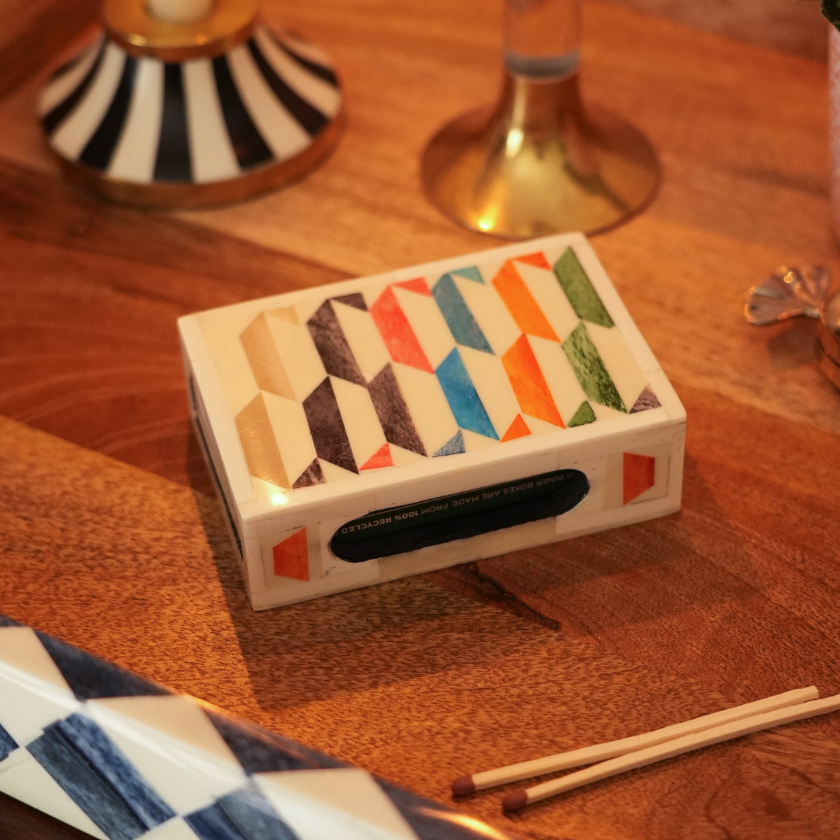 A decorative rectangular matchbox with a geometric multicolor pattern on top, placed on a wooden tabletop alongside matchsticks and other home decor items.