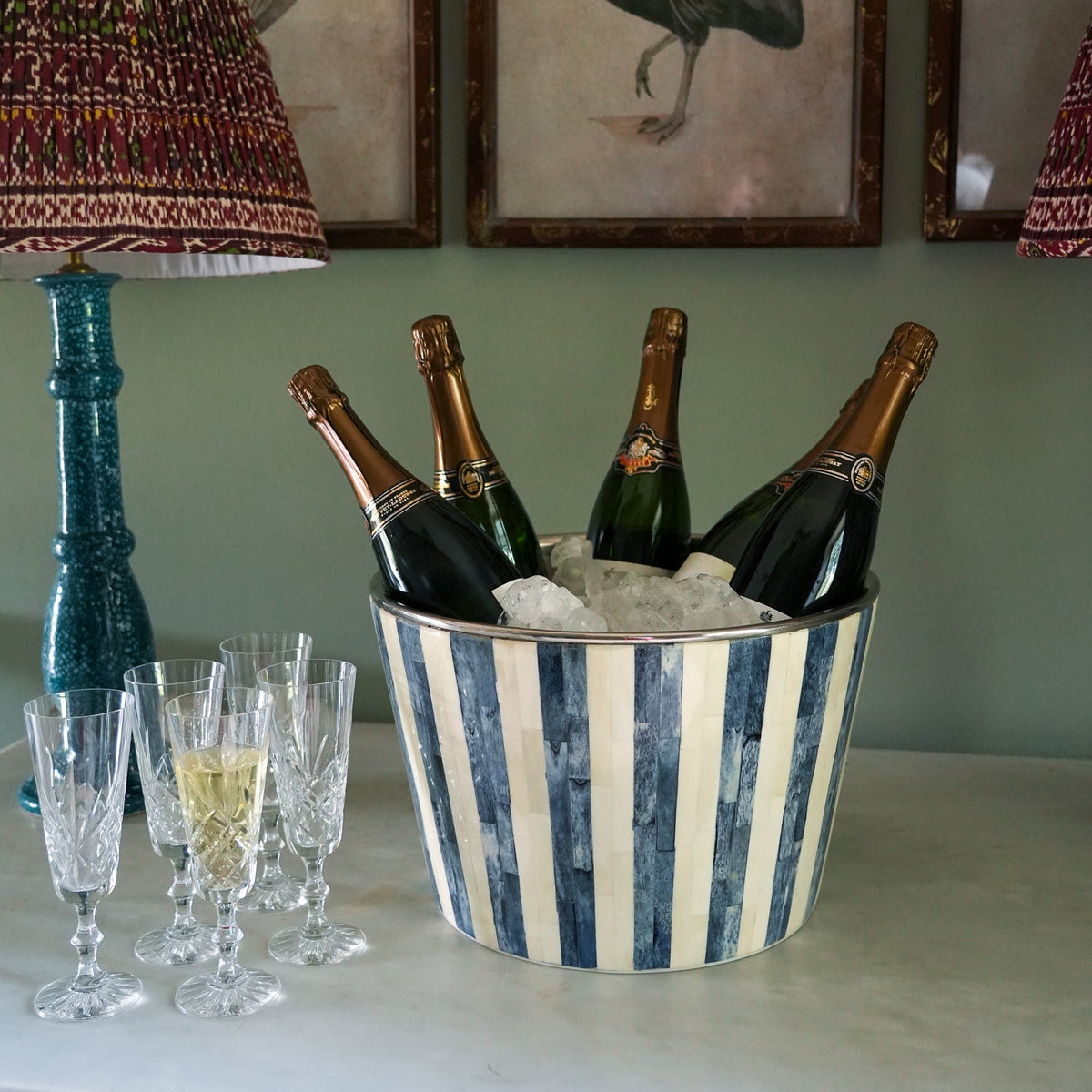 A blue and cream striped metal champagne bucket filled with ice and several champagne bottles, styled on a tabletop next to clear drinking glasses and a blue-green lamp in an indoor setting.