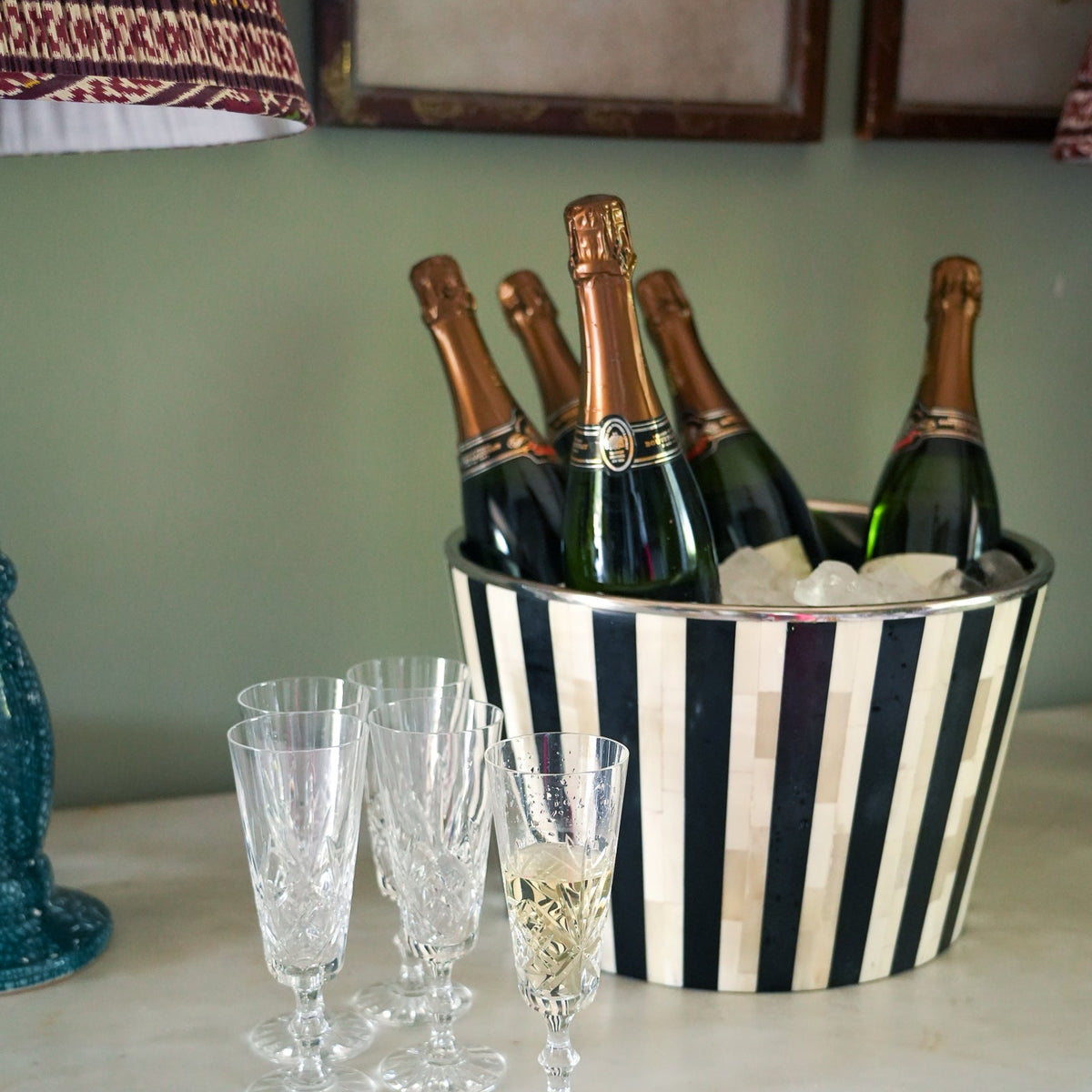 A black and white striped champagne bucket filled with ice and several champagne bottles, set on a white marble countertop next to four clear crystal champagne flutes.