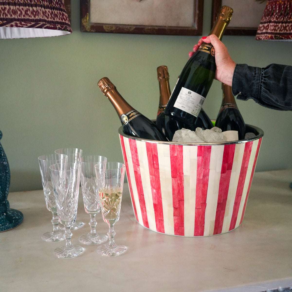 A red and white striped metal ice bucket filled with ice and several champagne bottles, with a hand holding one bottle. Three clear champagne flutes sit on the table beside it, with a green wall and framed art in the background.