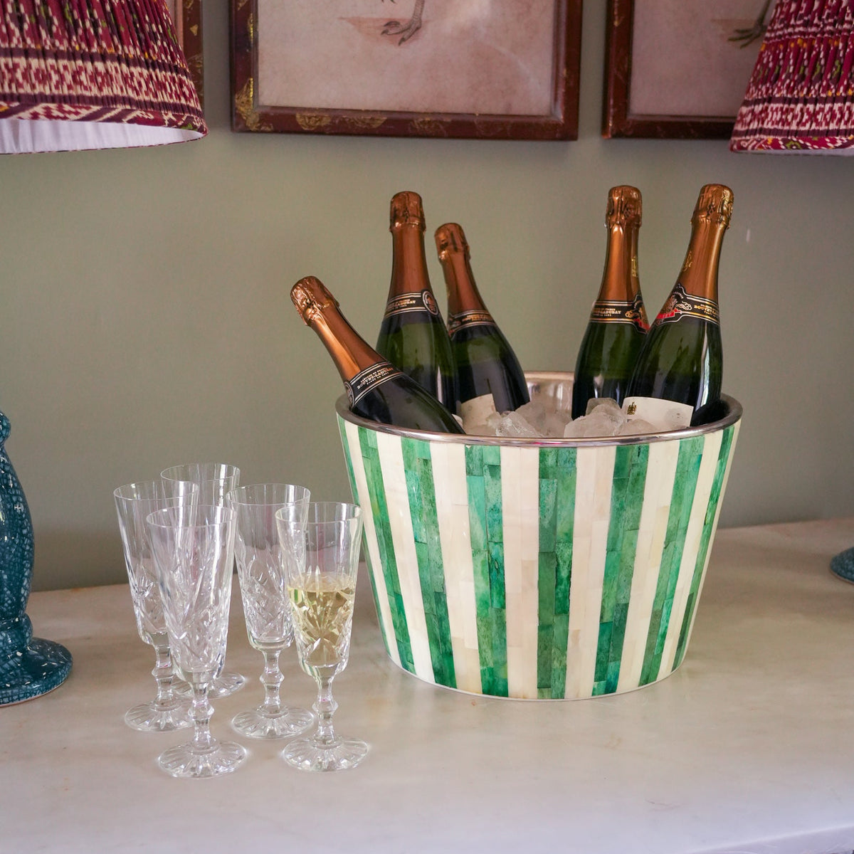 A green and white striped metal bucket filled with ice and several champagne bottles, placed on a white table next to four clear champagne flutes in an indoor setting.