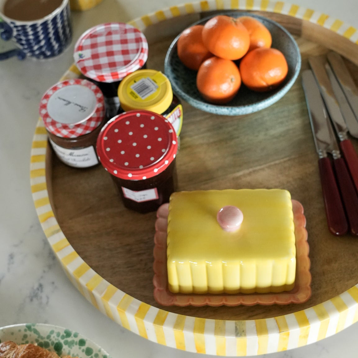 Wooden tray with butter, jam, and oranges on a checkered tablecloth.