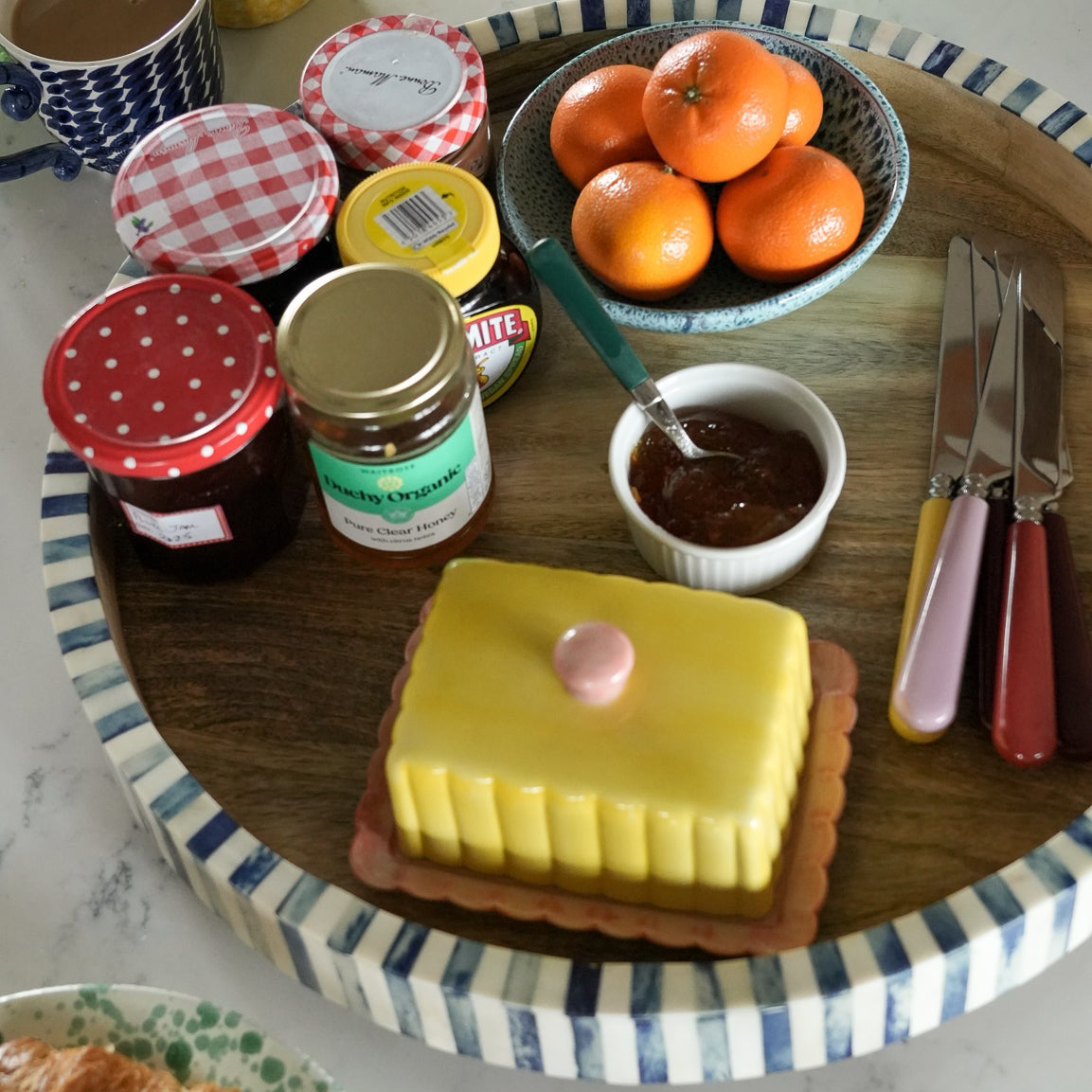 Table setting with a cake, jars of jam, oranges, and croissants on a decorative tray.