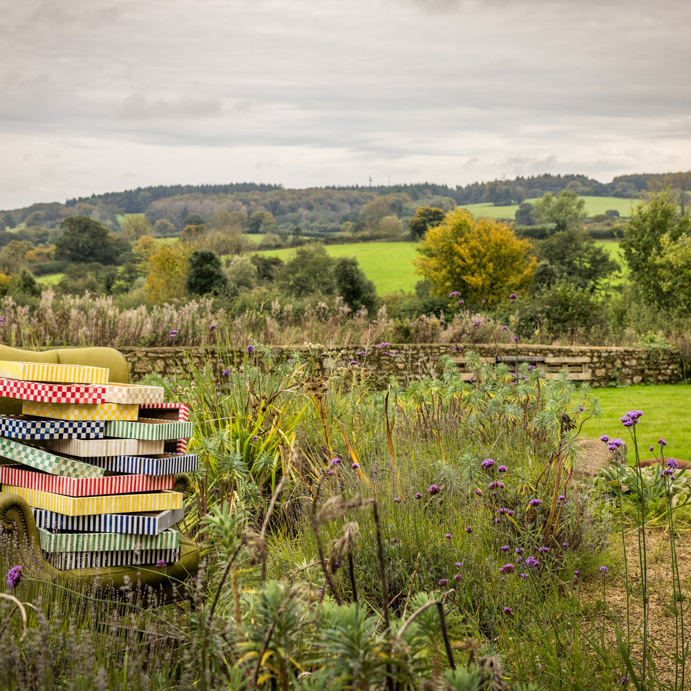 An English country garden landscape with olive green armchair amongst purple wildflowers and over a dozen colourful patterned wooden trays stacked on each other on the seat of the chair..