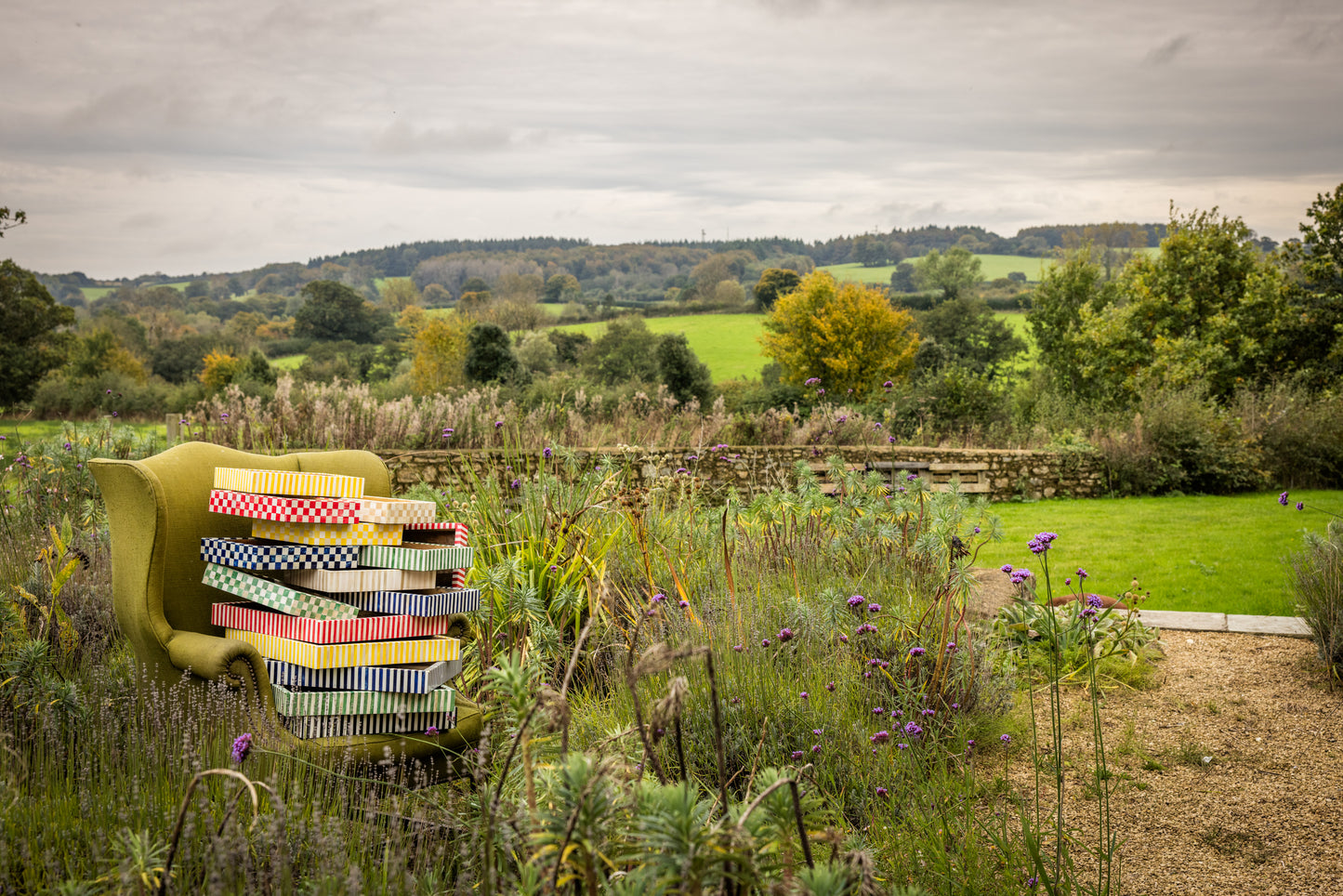 An English country garden landscape with olive green armchair amongst purple wildflowers and over a dozen colourful patterned wooden trays stacked on each other on the seat of the chair..